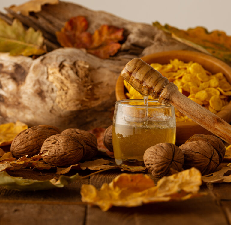 A wooden spoon with dripping honey on a jar, nuts and a bowl of cereal on autumn leaves blurry background